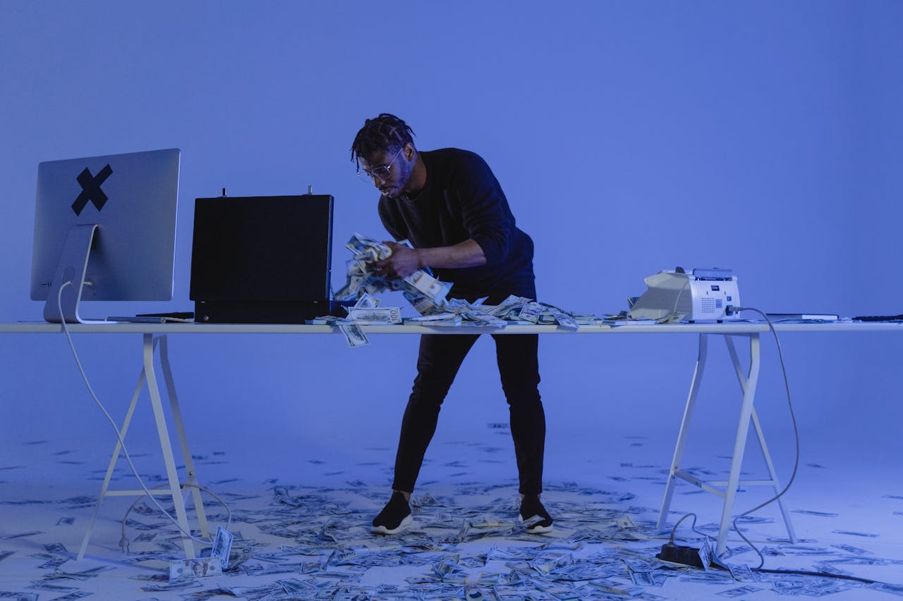 A man counting cash surrounded by computers and money in a blue-lit room, symbolizing cybercrime.